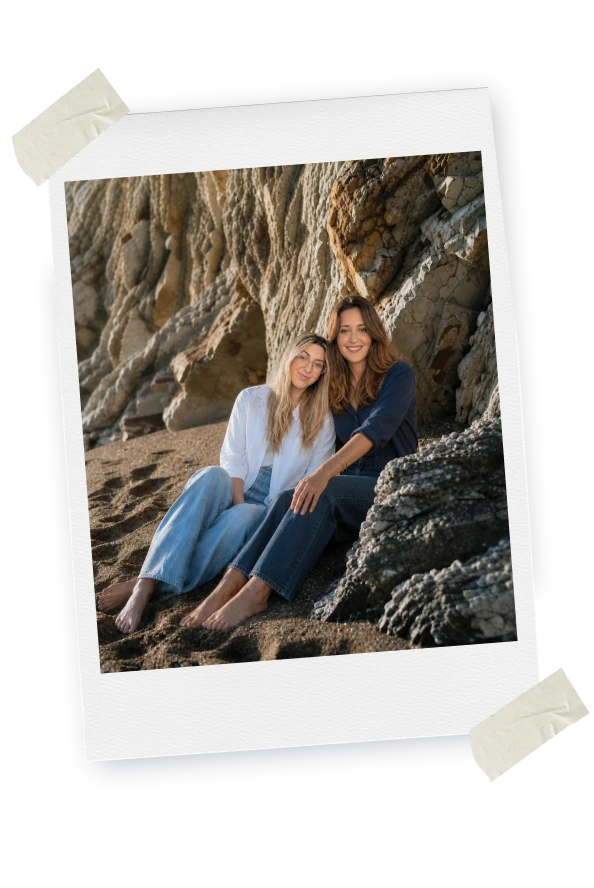 Cristina y Anabel sentadas en la playa junto a unas rocas.